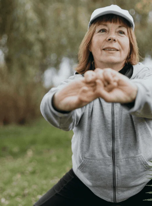 Older woman in a cap stretching her arms forward outdoors on a bright day