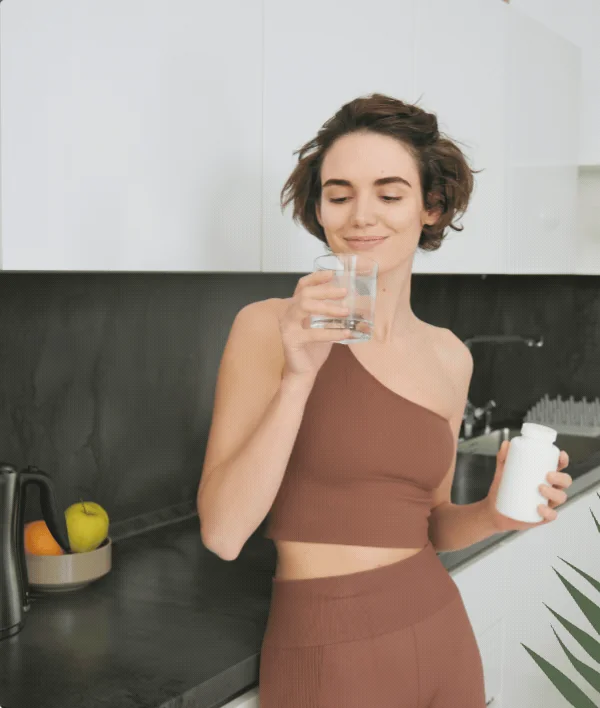 Woman drinking a glass of water in a bright kitchen beside a bowl of fresh fruit