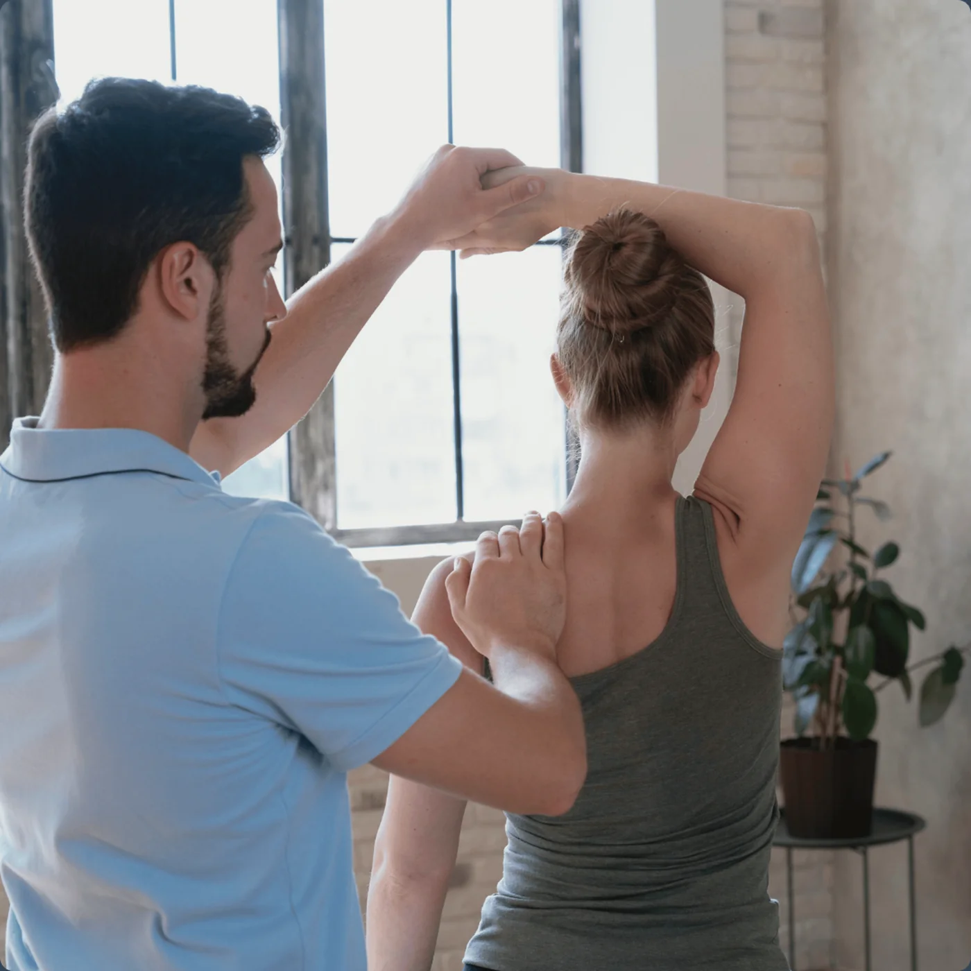 Practitioner guiding a standing patient through a shoulder and upper-back alignment check by a window